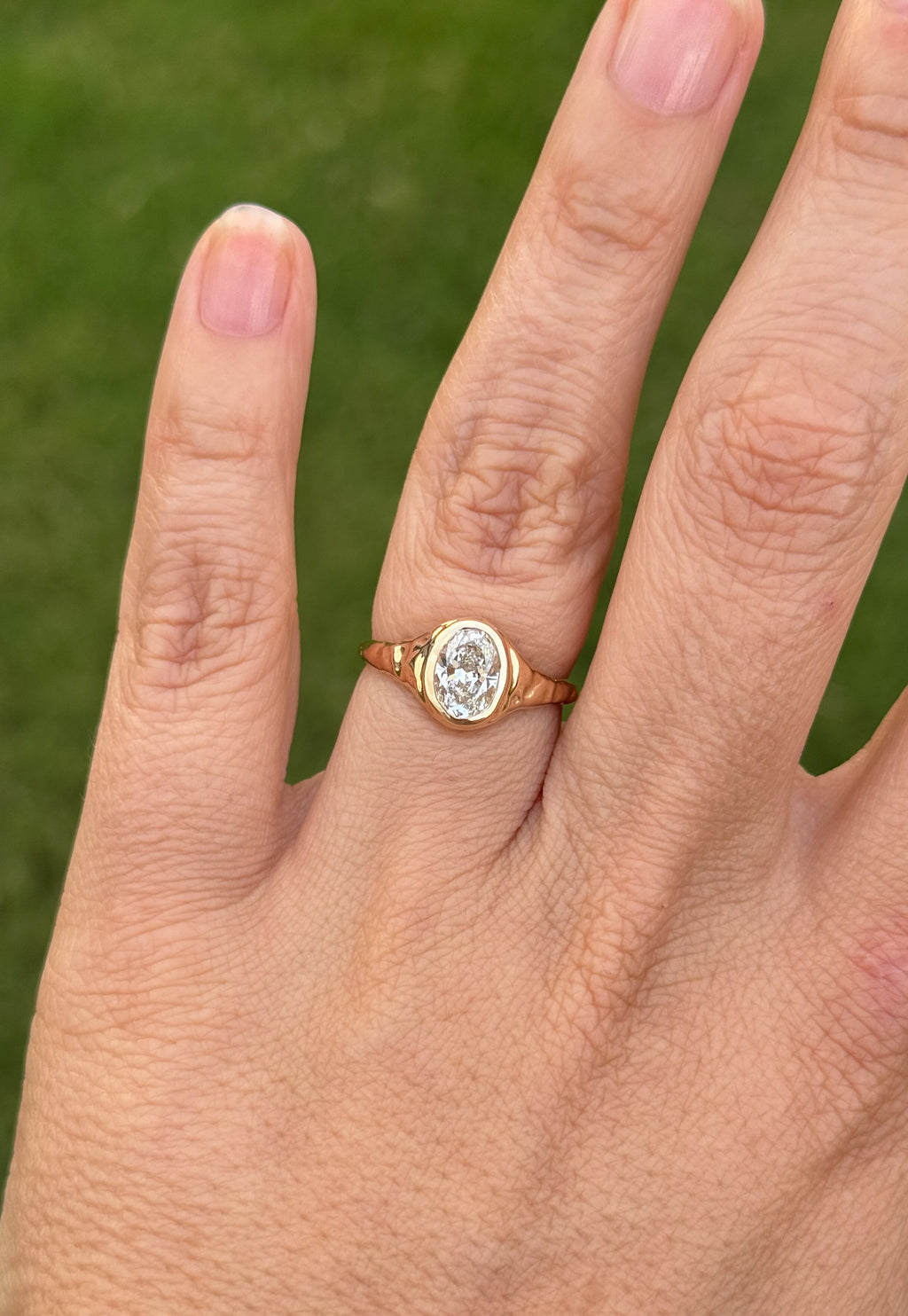 A close-up of a hand wearing a yellow gold ring with an oval-cut clear lab diamond, set against a blurred green background.