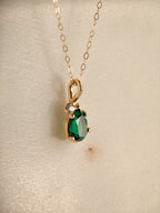 A gold pendant necklace featuring a faceted pear shape green emerald  gemstone and a small white sapphire accent stone, displayed on a textured beige surface.