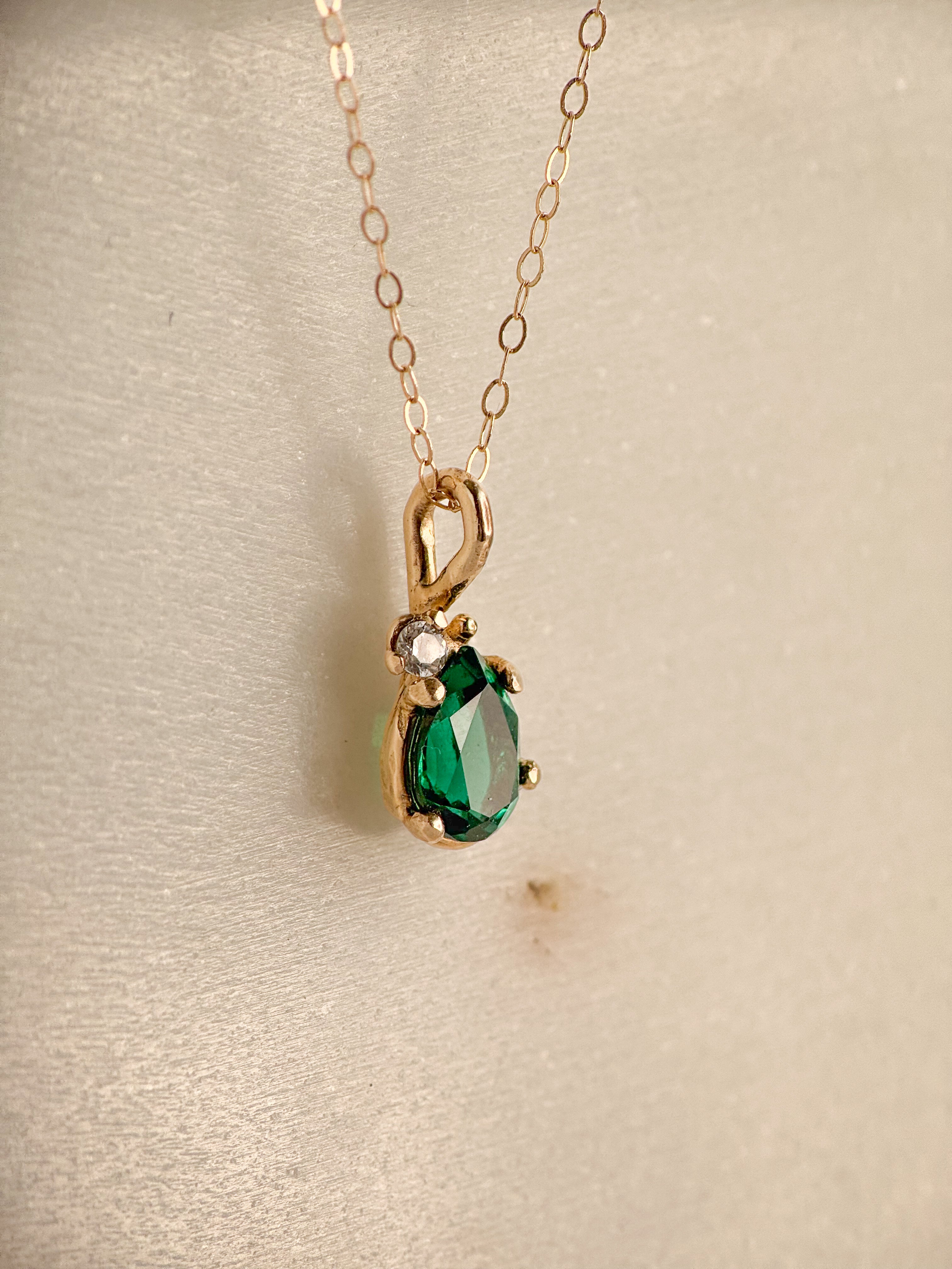 A gold pendant necklace featuring a faceted pear shape green emerald  gemstone and a small white sapphire accent stone, displayed on a textured beige surface.