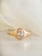 A gold ring with a textured molten band and an oval lab diamond, displayed on a soft cream-colored fabric background.