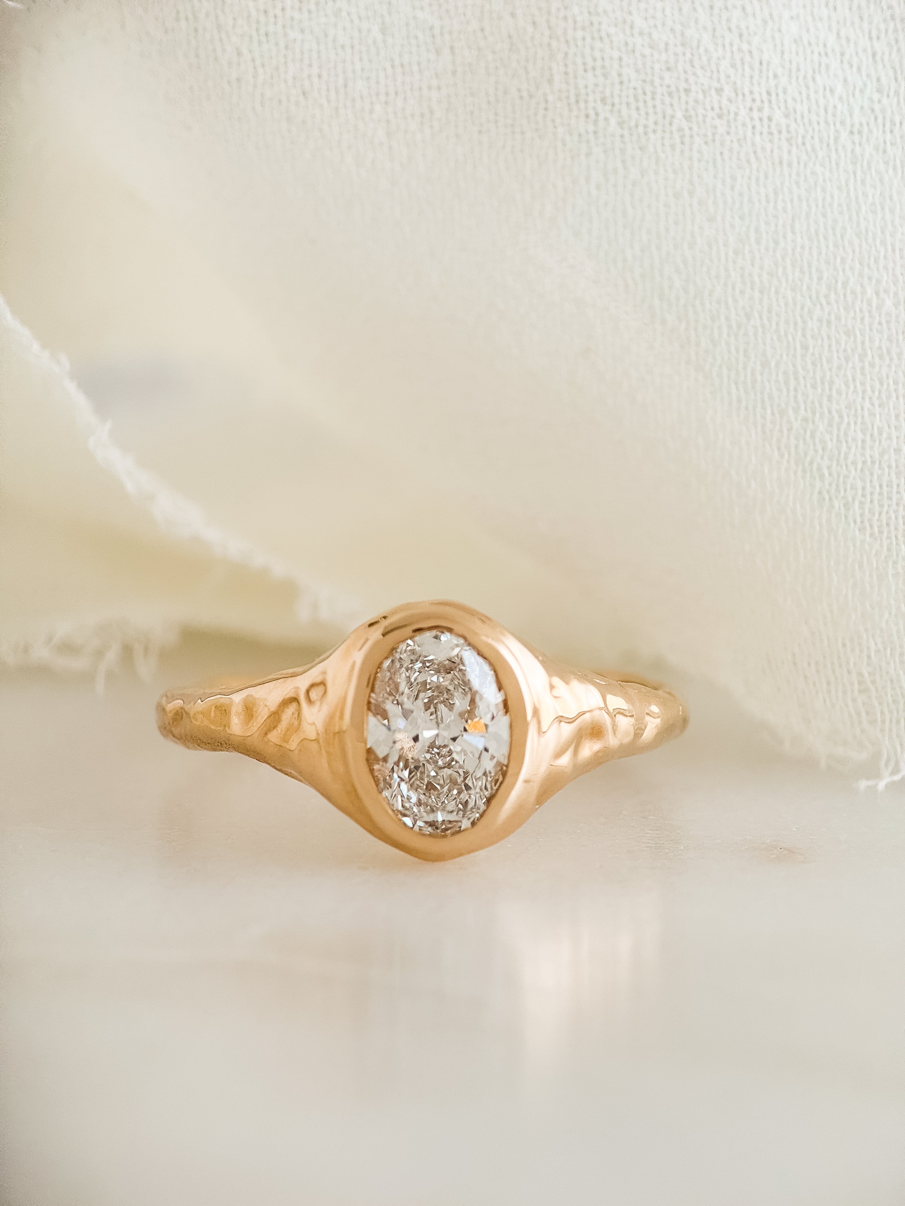 A gold ring with a textured molten band and an oval lab diamond, displayed on a soft cream-colored fabric background.