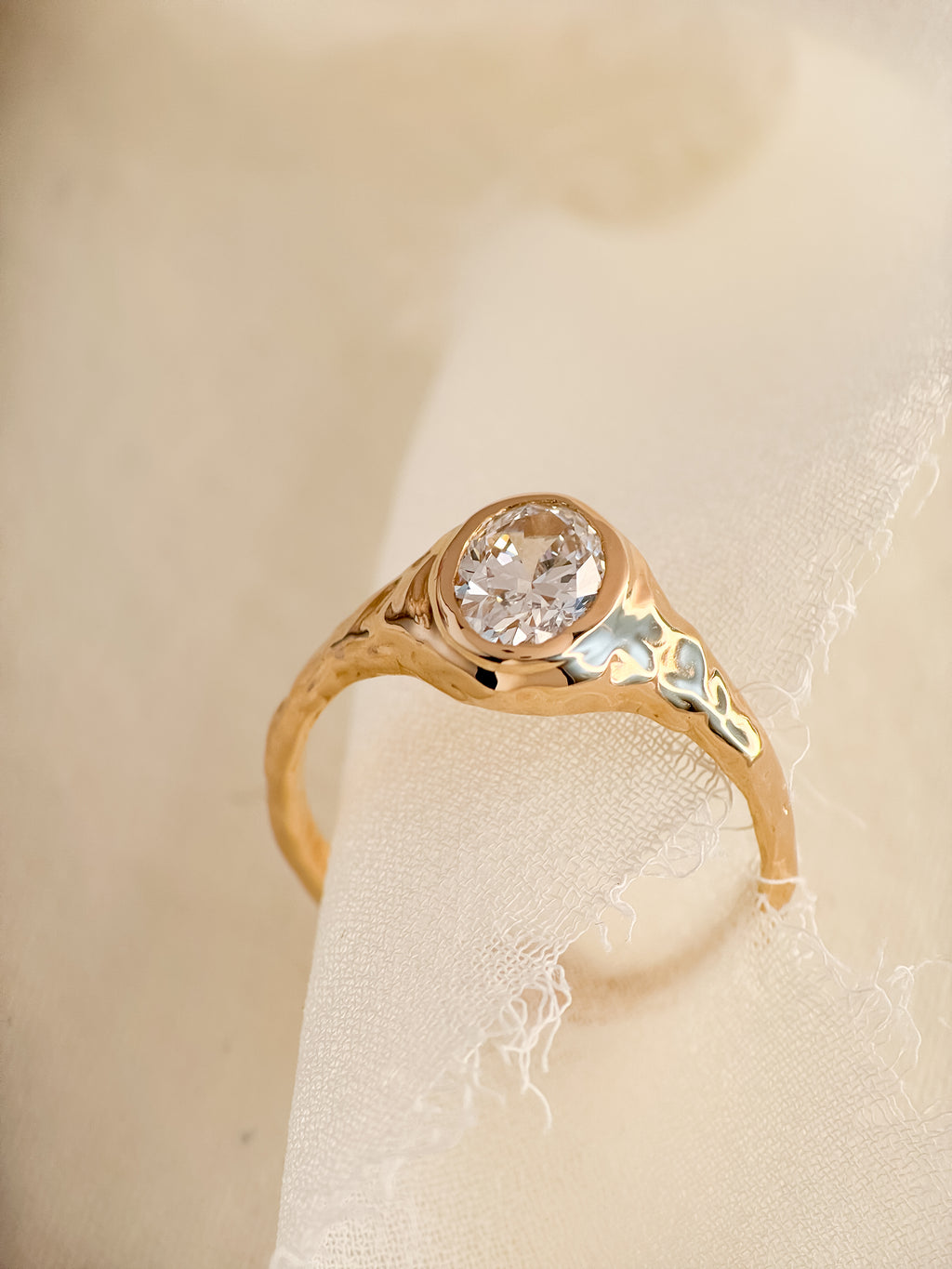 A gold ring with an oval lab diamondand textured, organic-shaped setting, displayed on a soft cream-colored fabric surface.