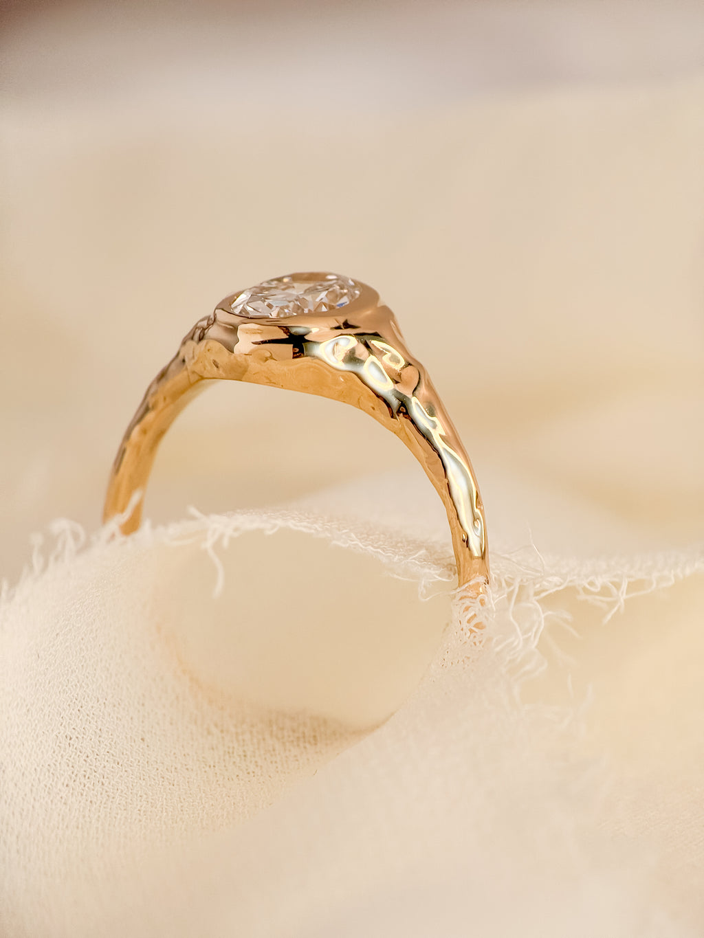 A gold ring with a textured, molten band and an oval lab diamond, resting on a soft beige fabric surface.