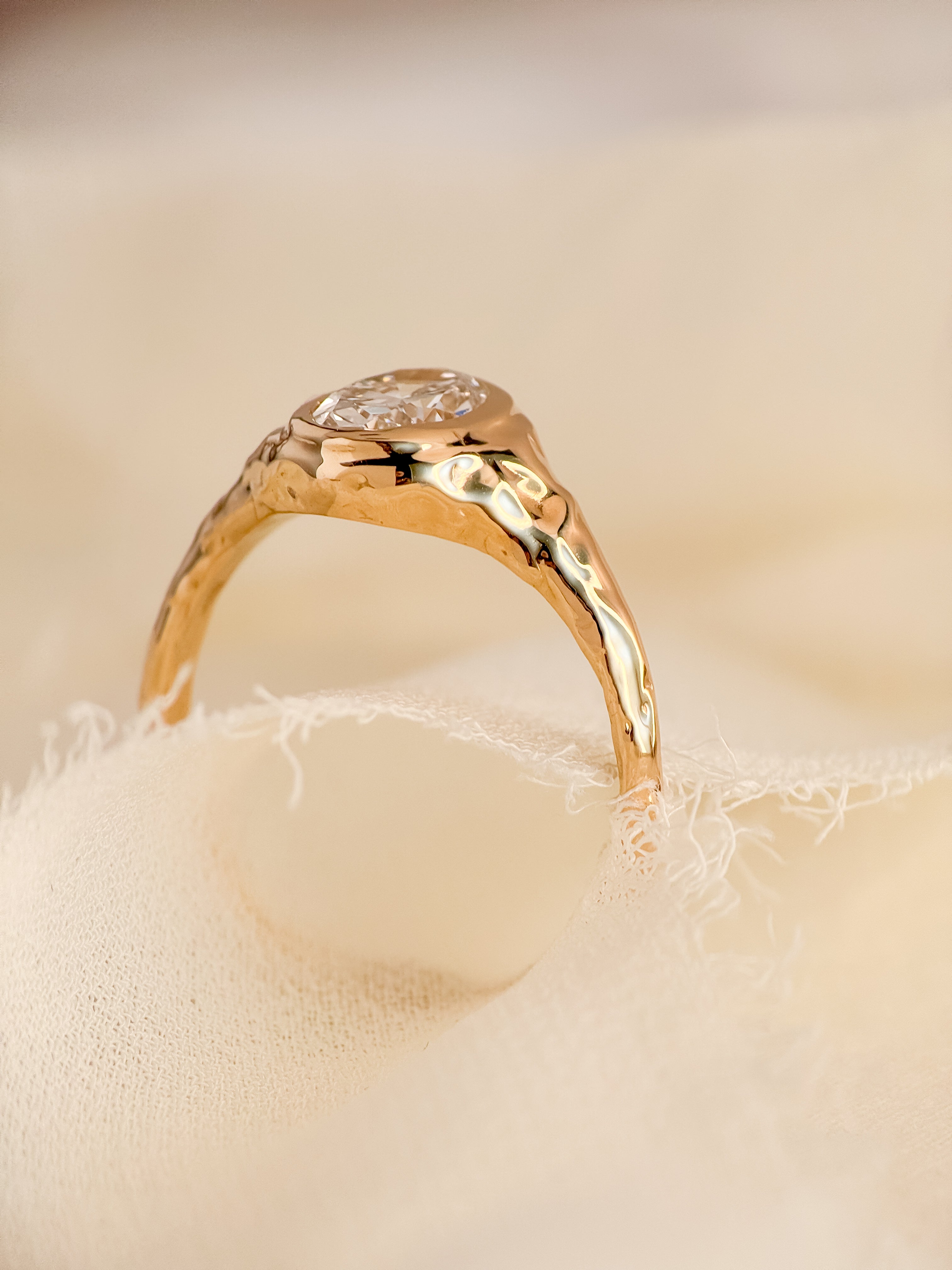 A gold ring with a textured, molten band and an oval lab diamond, resting on a soft beige fabric surface.