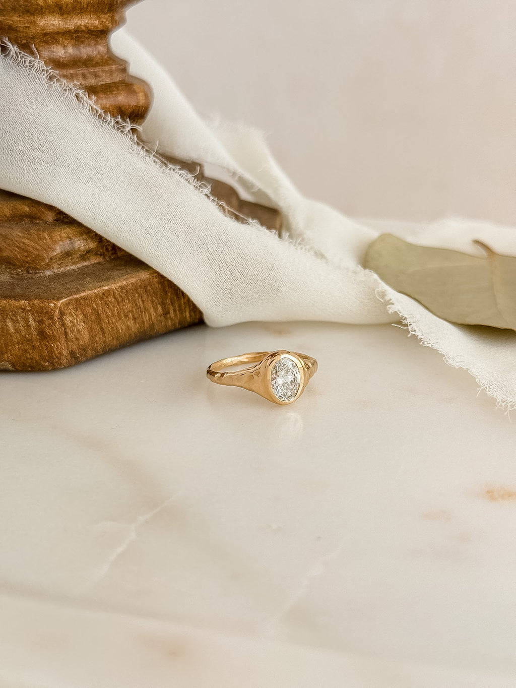 A gold ring with an oval diamond gemstone resting on a light marble surface, styled with a white fabric and a wooden block.