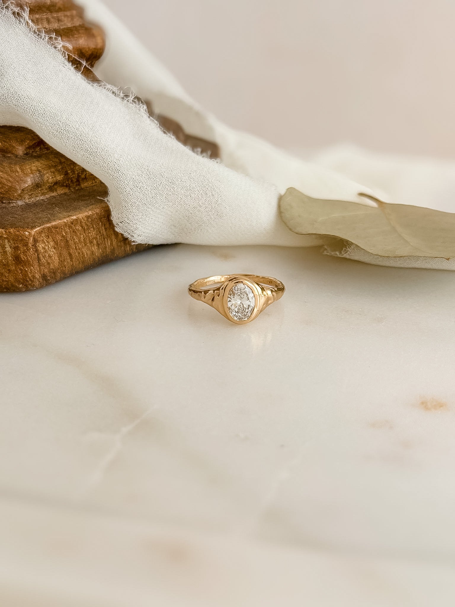 A gold ring with a bezel-set oval diamond gemstone, resting on a light marble surface with draped white fabric and a wooden block in the background.