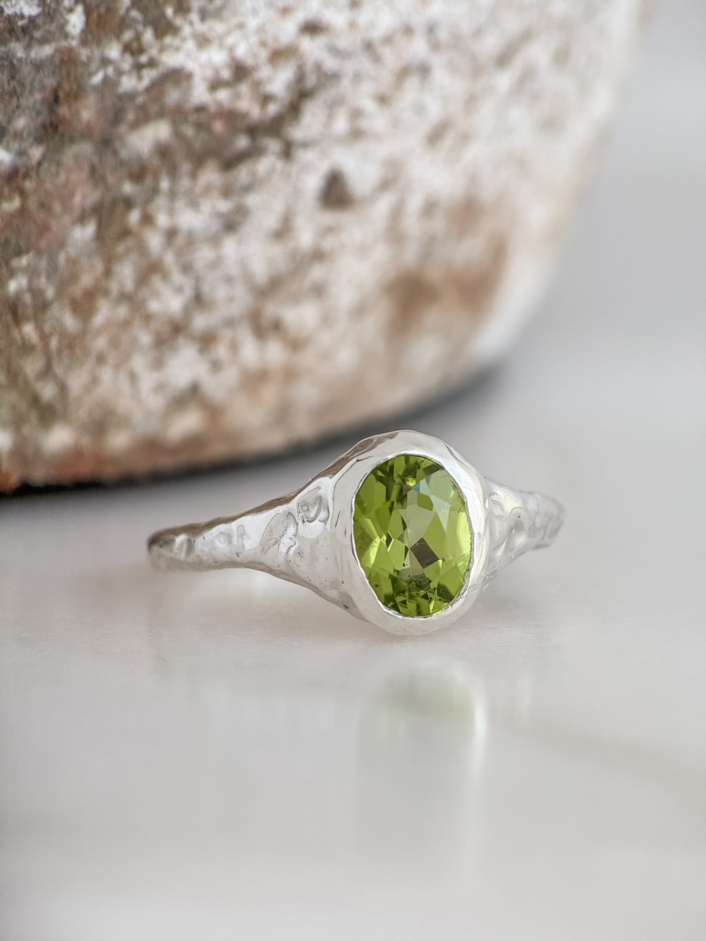 A sterling silver ring with a textured band and an oval-cut green peridot gemstone, resting on a light marble surface with a blurred stone in the background.