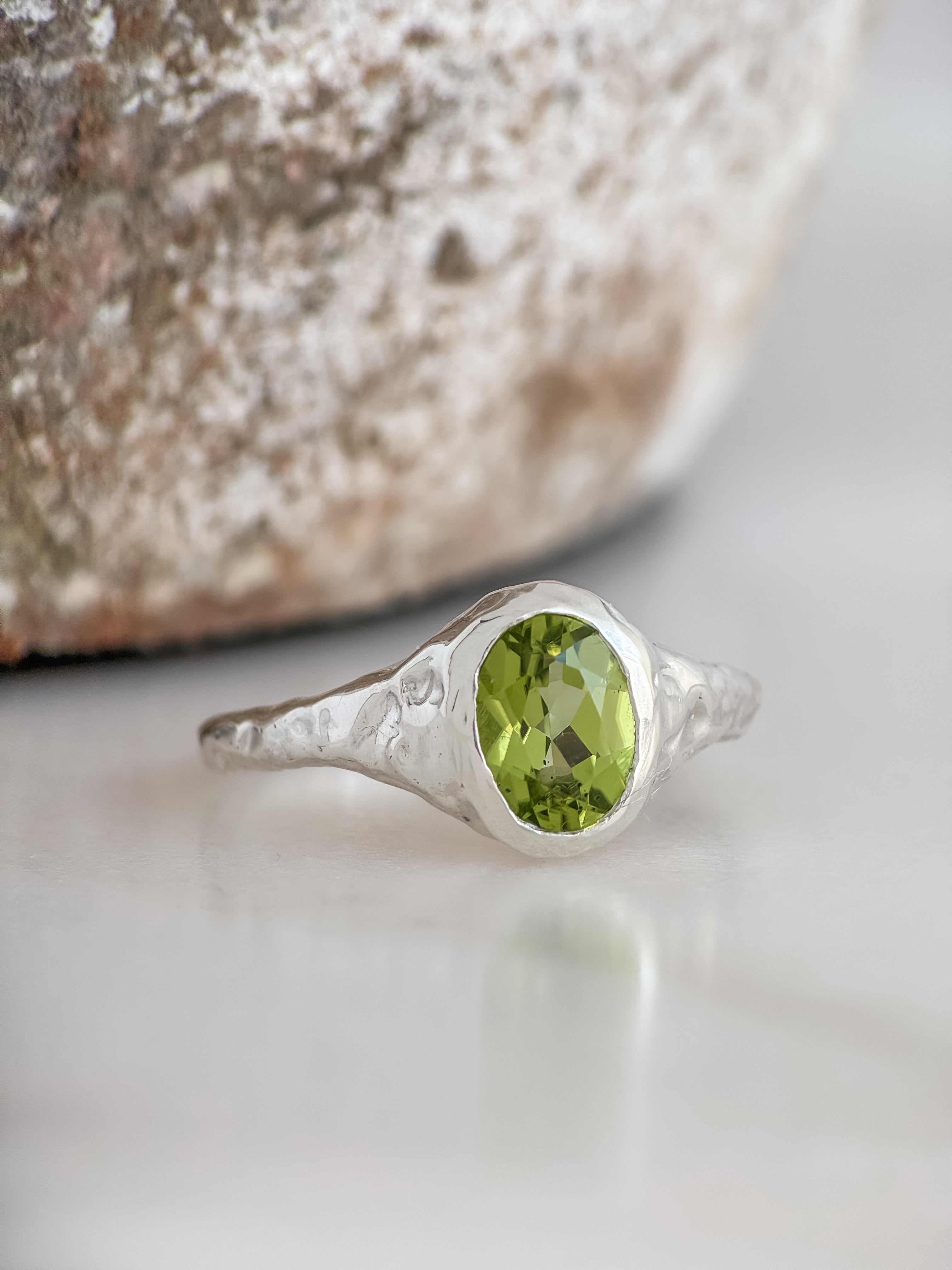 A sterling silver ring with a textured band and an oval-cut green peridot gemstone, resting on a light marble surface with a blurred stone in the background.