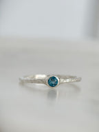 A silver ring with a textured band and a round London blue topaz gemstone in a bezel setting, resting on a light gray reflective surface.