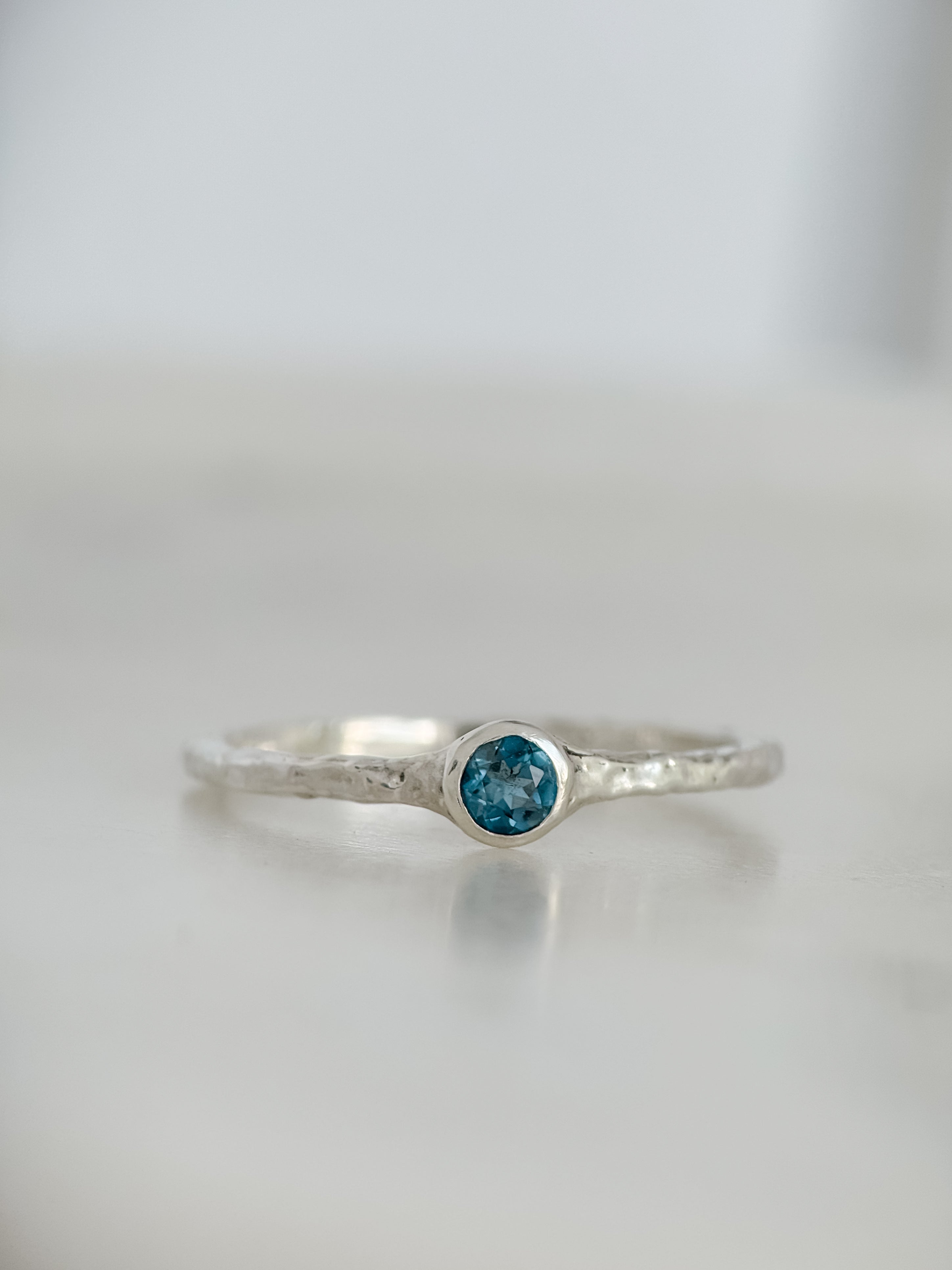 A silver ring with a textured band and a round London blue topaz gemstone in a bezel setting, resting on a light gray reflective surface.
