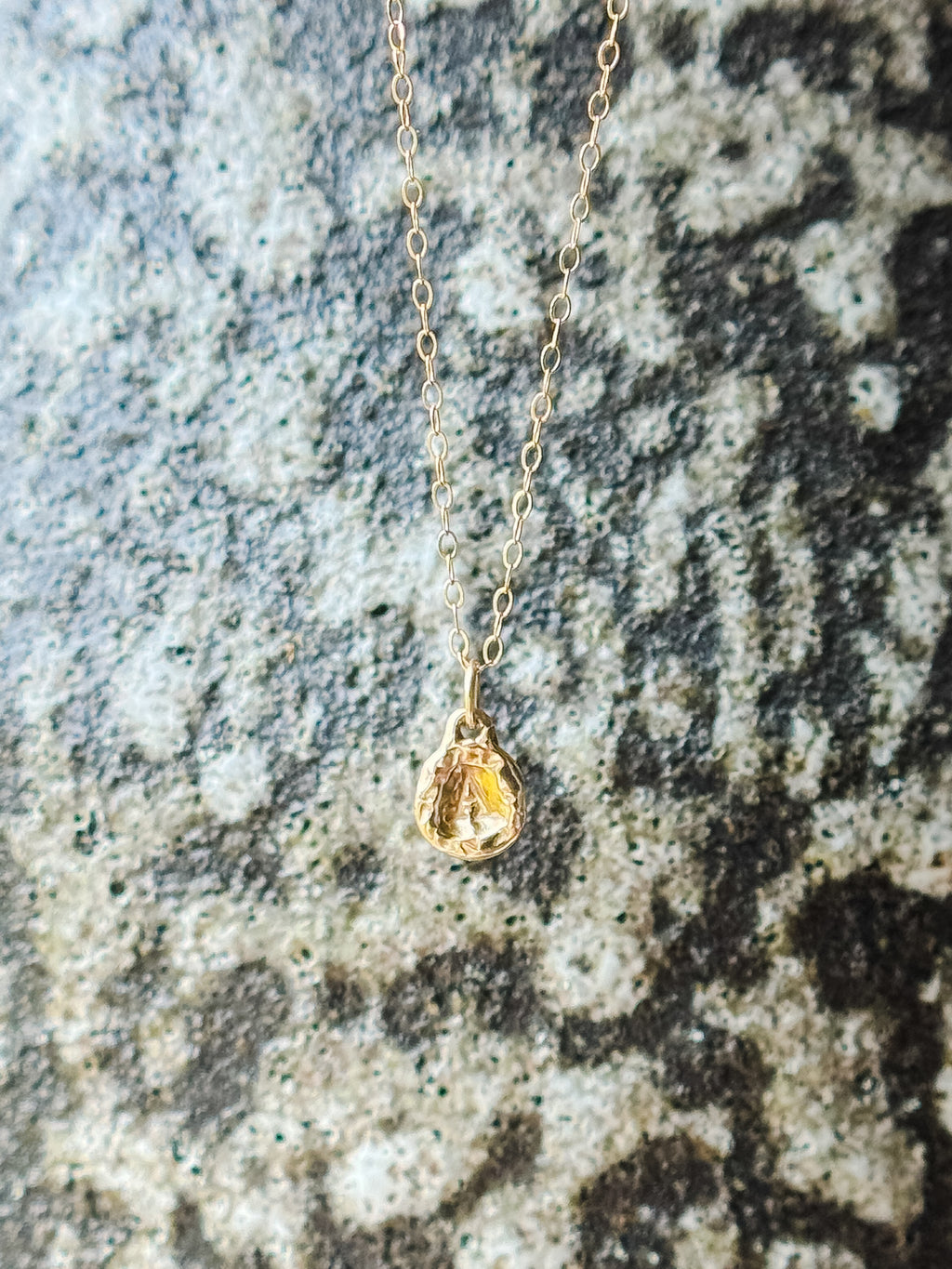 A delicate gold chain necklace with a small molten initial texture, resting on a textured gray and white stone surface.