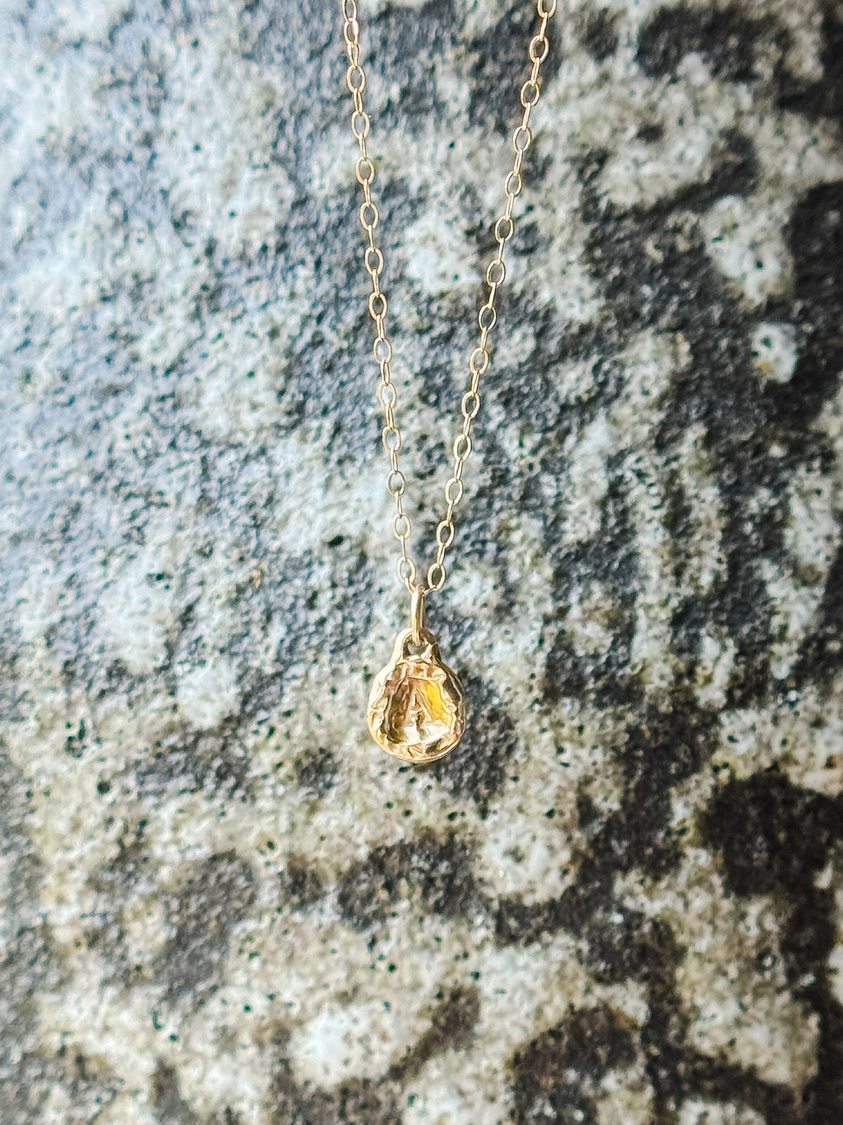 A delicate gold chain necklace with a small molten initial texture, resting on a textured gray and white stone surface.