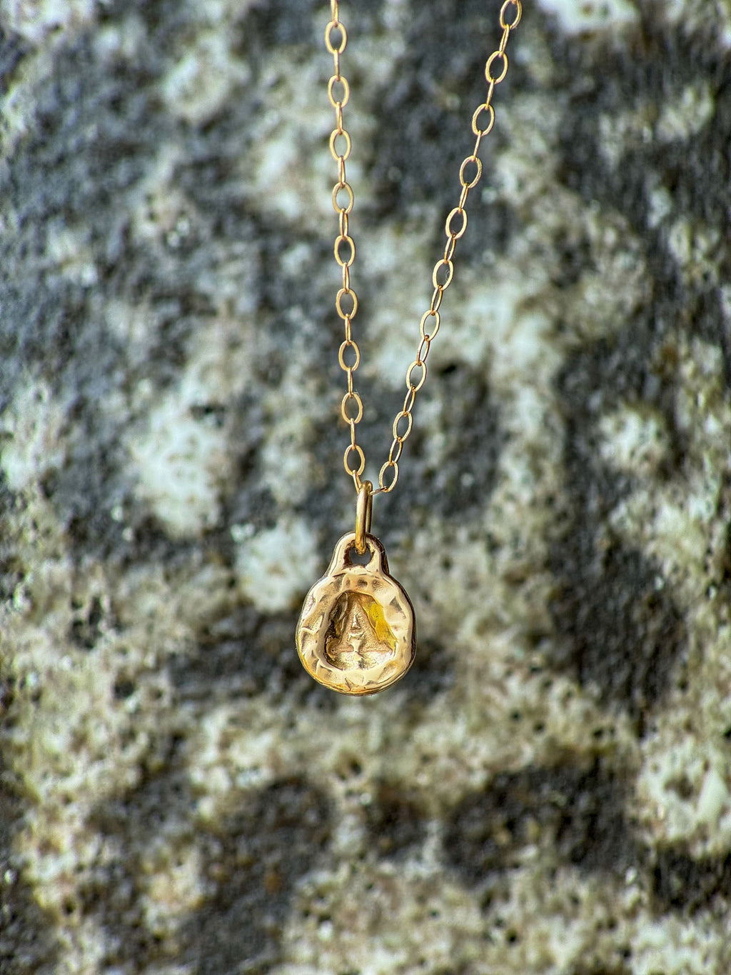 A delicate gold chain necklace with a small molten initial texture, resting on a textured gray and white stone surface.