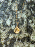 A delicate gold chain necklace with a small molten initial texture, resting on a textured gray and white stone surface.