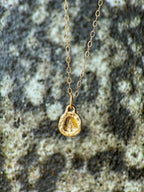 A delicate gold chain necklace with a small molten initial texture, resting on a textured gray and white stone surface.