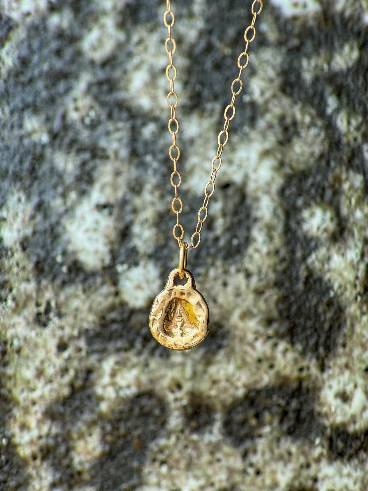 A delicate gold chain necklace with a small molten initial texture, resting on a textured gray and white stone surface.