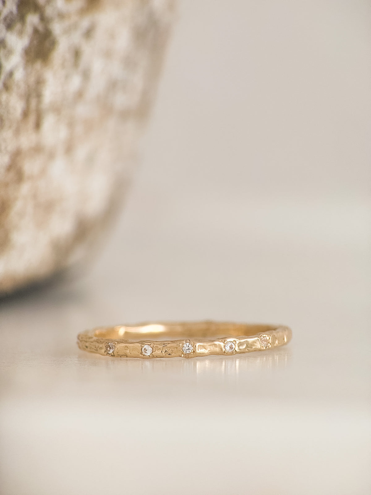 A thin yellow gold band ring with a textured molten finish and a row of small diamonds, resting on a light beige surface with a soft-focus decorative object in the background.