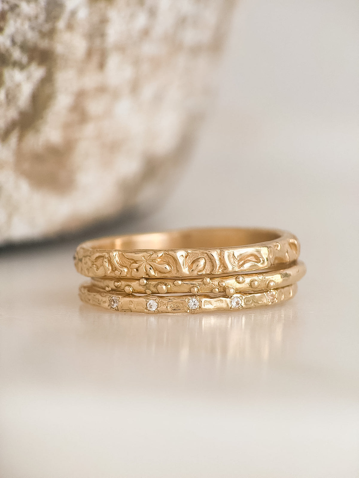 Three stacked gold rings with textured, engraved patterns and small clear diamonds, displayed on a light surface with a soft-focus decorative background.