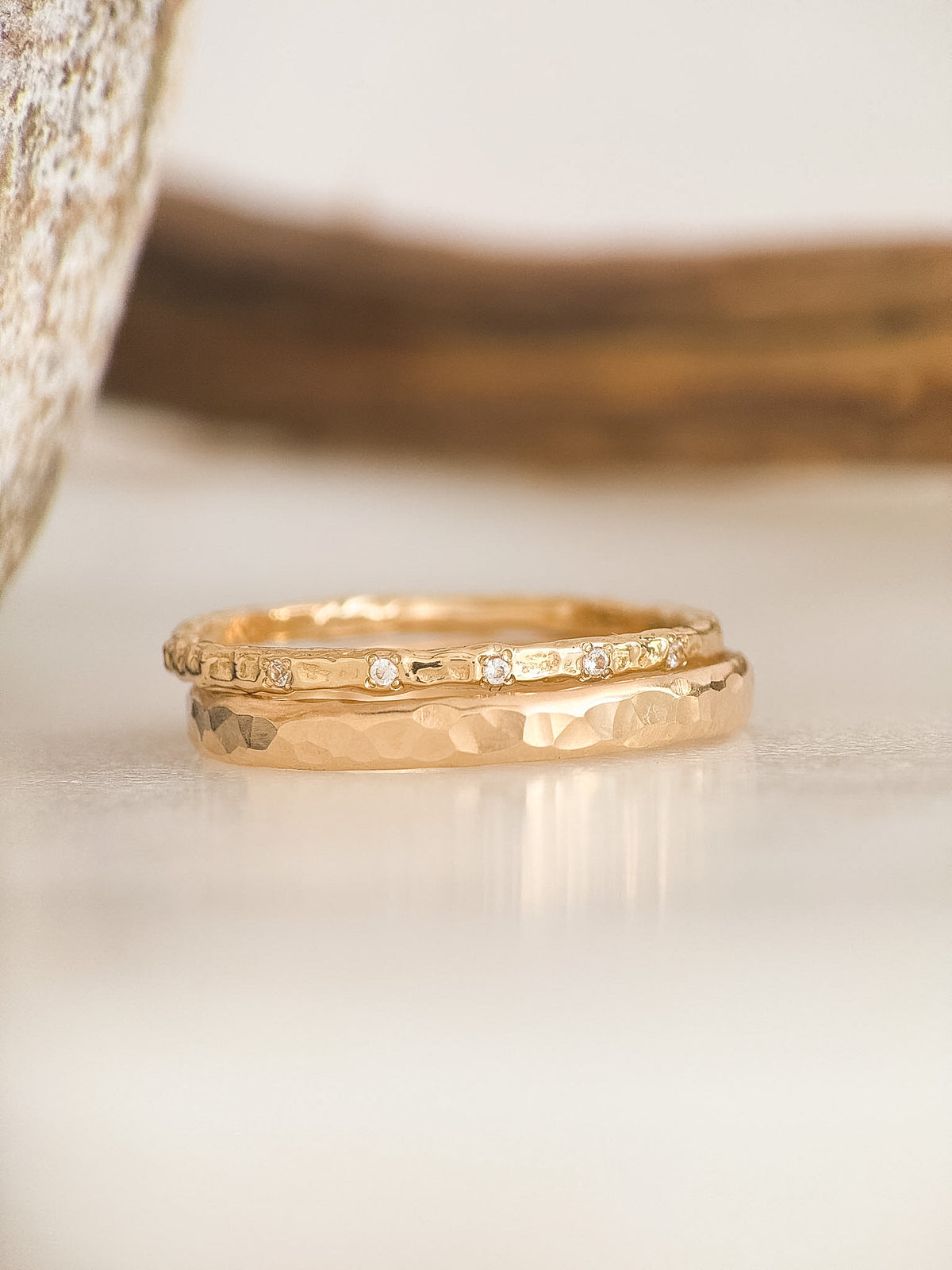 Two gold rings, one with a hammered texture and one with a molten finish and small diamonds, displayed on a light surface with a blurred background.
