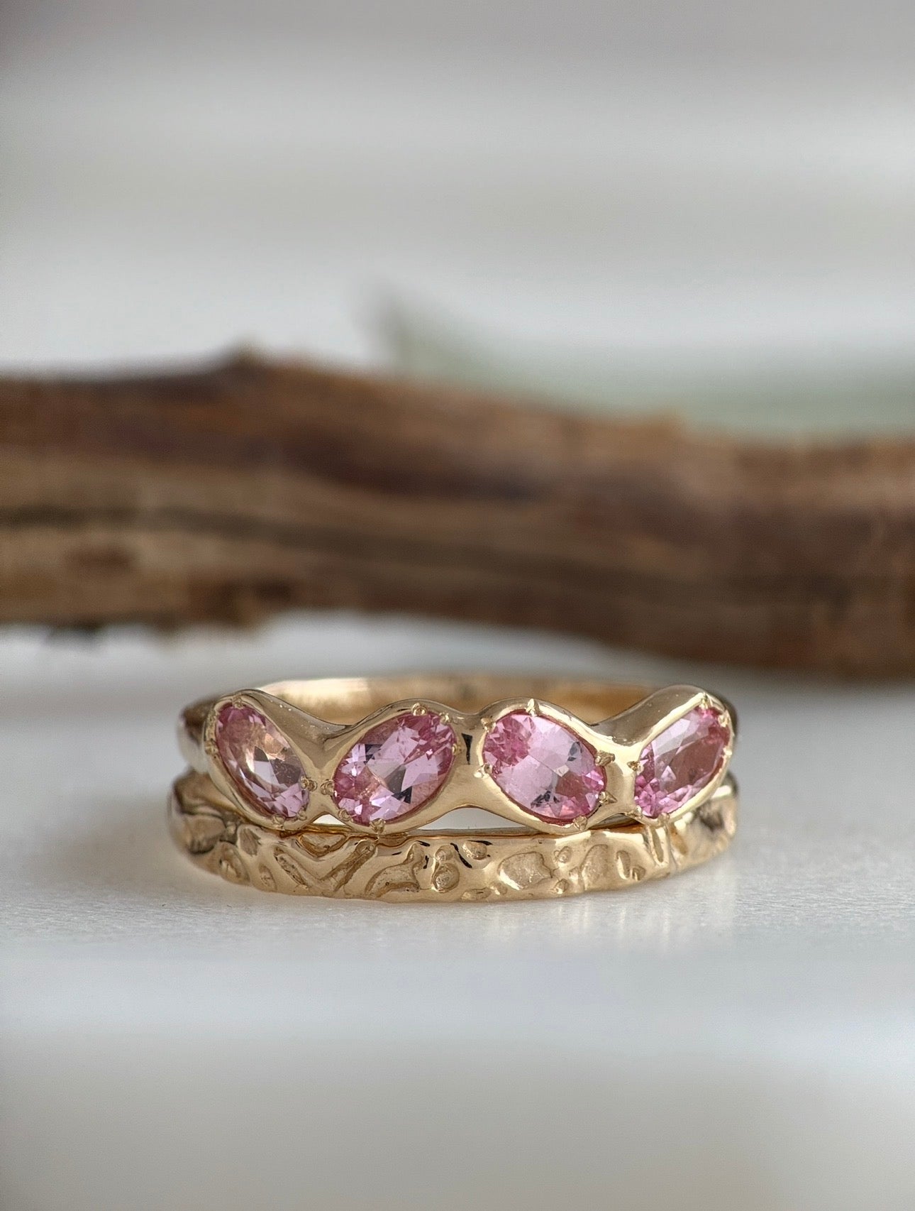 Gold ring with pink tourmaline gemstones stacked on top of a textured gold ring on a white surface