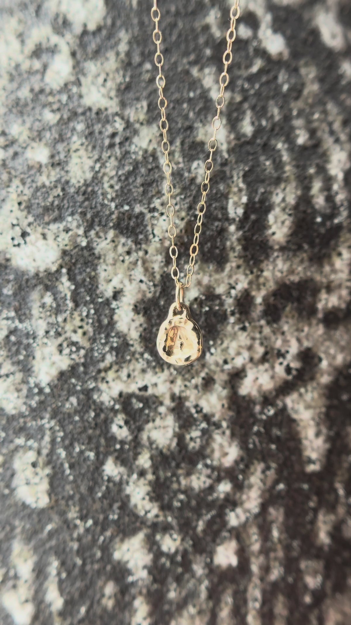 Pebble Initial Charm Necklace in 14K yellow gold dangling in front of a white and gray spotted background.